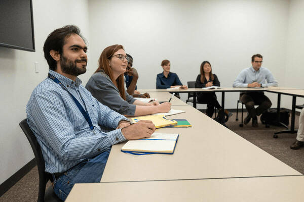 Bearded man in blue striped shirt smiles, writing in a notebook at a classroom table with diverse students.