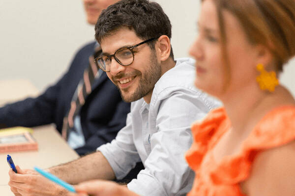 Smiling man with beard, glasses, and light blue shirt holds a blue pen. Others blurred in background.