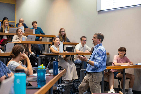 Professor in blue plaid shirt gestures while speaking to students with laptops in a tiered Notre Dame lecture hall.