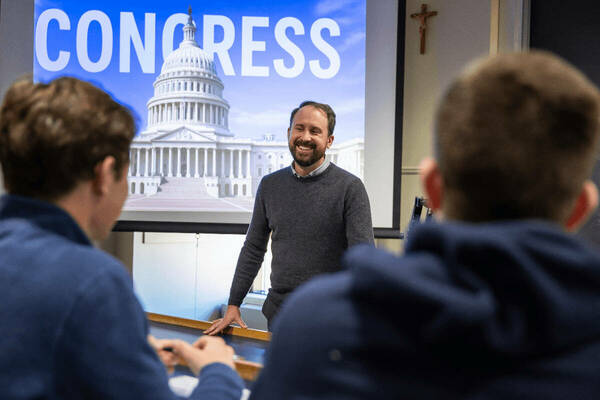 Smiling bearded man in gray sweater presents. Behind him, a screen displays the US Capitol and "CONGRESS" on blue.