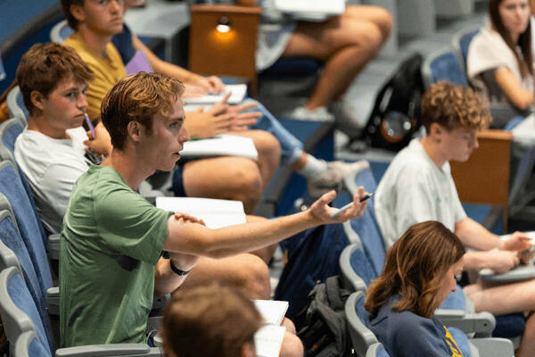 A male student in a green shirt extends his arm, holding a pen, while speaking in a lecture hall with other students.
