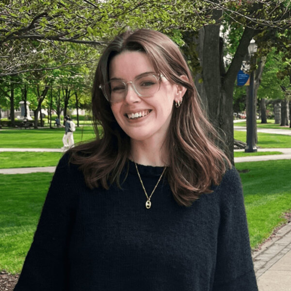 A young woman with brown hair, clear glasses, and a black sweater smiles broadly on the Notre Dame campus with lush trees.
